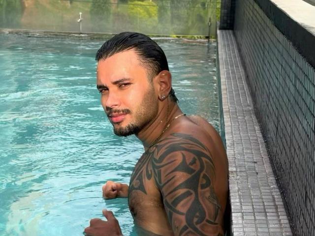 Aletargaryem, con una spalla tatuata, capelli neri bagnati e una catena d'oro, è in piedi in una piscina con piastrelle blu, guardando indietro con un leggero sorriso. Il suo sedere è parzialmente visibile..
