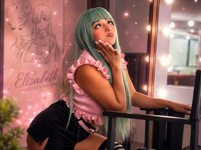 AilenRosse with long green hair, pouty lips, pink ruffled top, black shorts, and a pink belt, leaning on a mirror, looking up with a hand on her cheek.