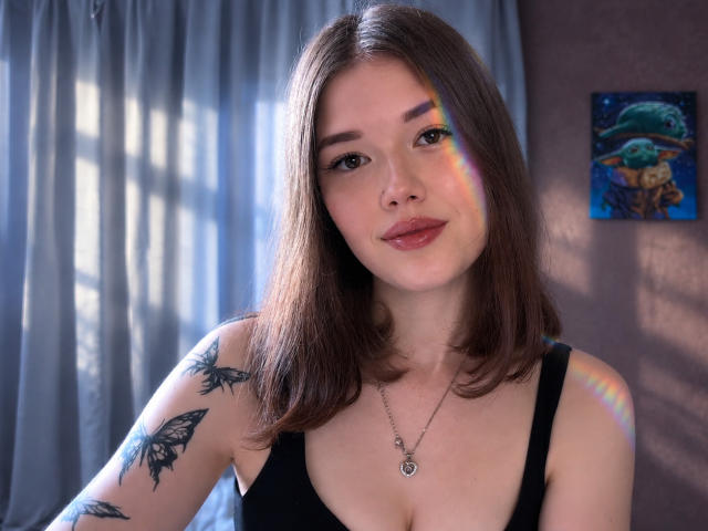 TrudieProse with shoulder-length brown hair, black tank top, butterfly tattoos, and a necklace, showing a subtle smile with rainbow reflection on her face.