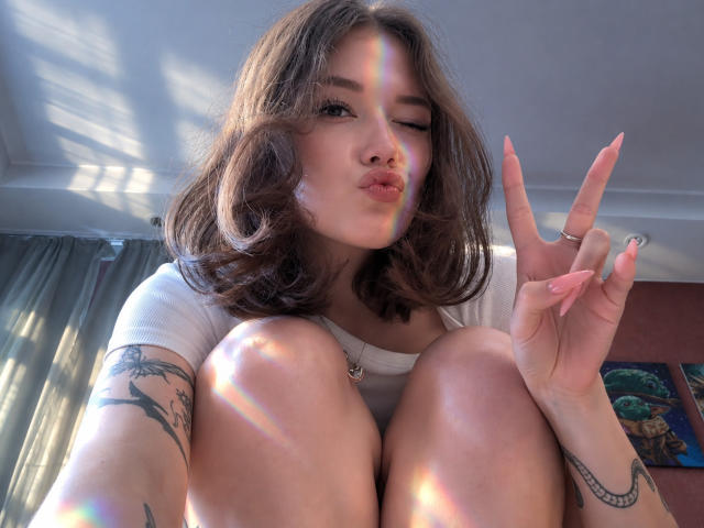 TrudieProse with wavy brown hair, white tee, and tattoos, squatting, making a pouty face, and flashing a peace sign, with a rainbow reflection on her face.