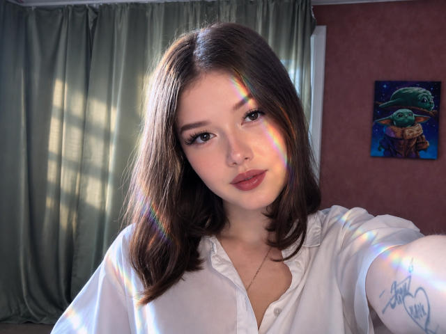 TrudieProse with medium brown hair, wearing a white shirt, has a rainbow light reflection on her face, a nose ring, and a tattoo on her arm. Soft lighting, cute.