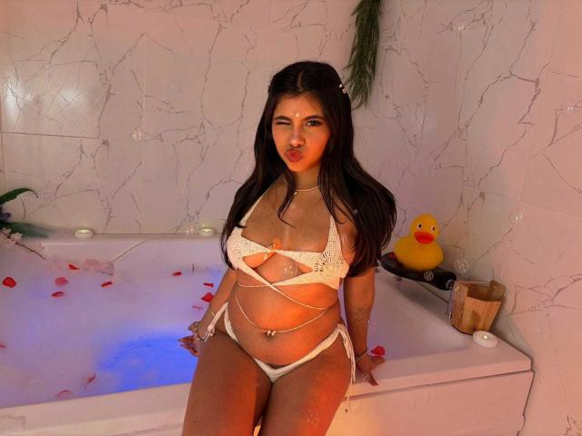 CelesteMorel with big tits, wearing white strappy lingerie, sitting in a bathtub filled with rose petals, her wet ass and belly button ring visible, and a yellow rubber duck.