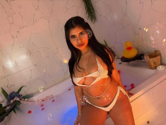 CelesteMorel with large tits, wearing a white lace bikini, standing in a bubble-filled bathtub, dark long hair, brown eyes, glowing face, yellow rubber duck, rose petals, and candles.