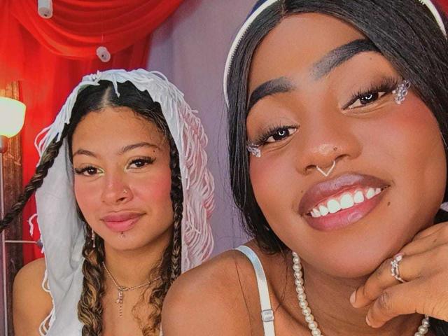 HaryAndKimmy, two dark-skinned women with braids, white veils, nose rings, and pearl necklaces, smiling, showing tits, with one's hand on her chin, in a selfie.