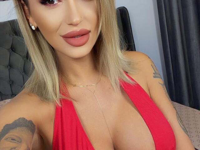 NicolSNaya, avec des cheveux blonds, un haut rouge à encolure américaine, de grandes seins, des tatouages, une chaîne en or, du rouge à lèvres rose, souriante, regardant la caméra..