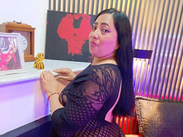 AmberMoretti with long black hair, black lace dress, and visible ass, leaning on a white counter, looking back with a slight smile, red bracelet, wooden clock, and gold figurine.