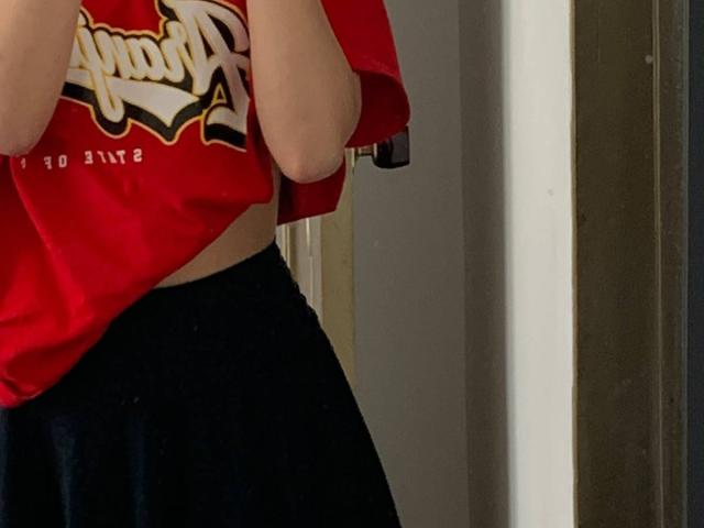 HannaThorne's red shirt lifted to reveal her belly, wearing black pants, her hands near her face, her shirt has gold and white lettering, no visible pussy or ass.