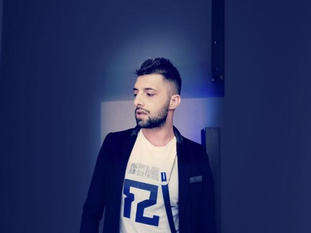 TommyDum with short, dark hair and beard, wearing a black blazer over a white shirt with blue number "22", standing and looking to the side with a serious expression.