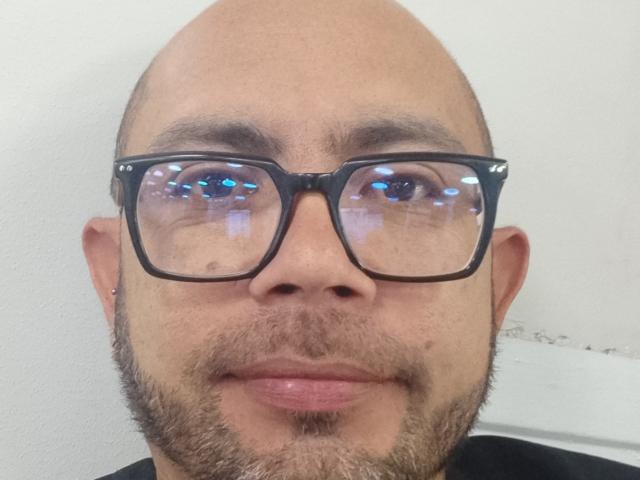 A cabeça careca de Kaelo e sua barba marrom, com óculos pretos, refletindo luzes azuis em seus olhos. Ele tem uma pequena pinta no nariz e uma pequena perfuração na orelha..
