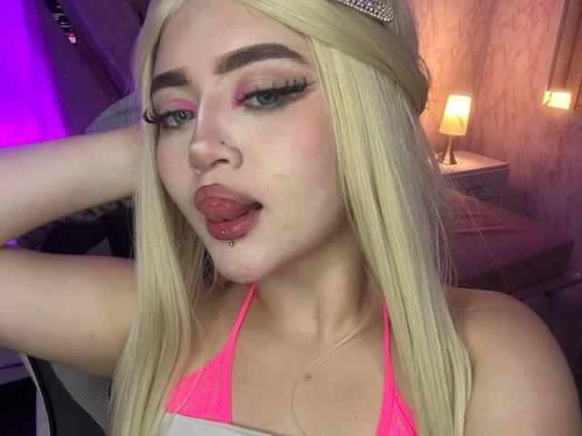 Lyanekkoo, avec ses cheveux blonds, ses paupières roses et sa langue percée, sort sa langue. Elle porte un haut de bikini rose néon et un anneau de nez. Elle a de longs cils et une petite piercing sur la lèvre..