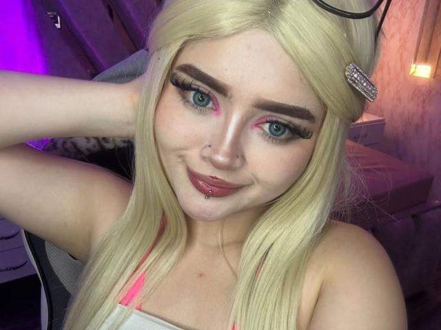 Lyanekkoo, con i capelli biondi, gli occhi azzurri, l'eyeliner rosa e un piercing al labbro, sorride leggermente con una mano dietro la testa, indossando una maglietta rosa..