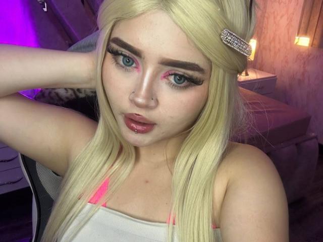 Blond Lyanekkoo with pink eyeliner, long eyelashes, and a pink lip piercing, wearing a white tank top, has a diamond hair clip, and a hand in her hair.