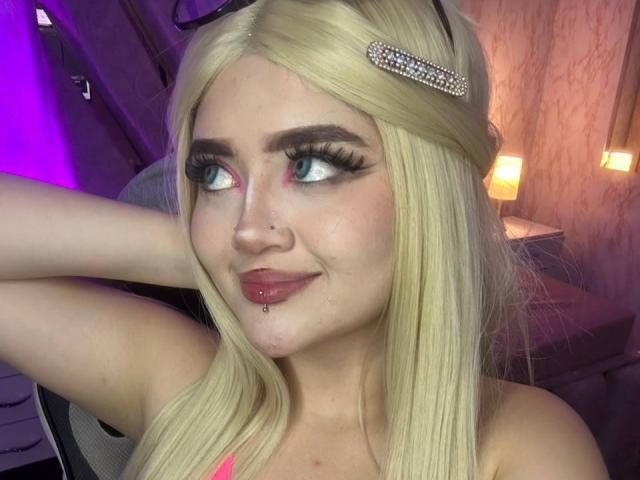 Lyanekkoo tem cabelo loiro, delineador rosa, cílios longos, um piercing no nariz, um piercing no lábio e um sorriso. Ela está usando um prendedor de cabelo brilhante e uma blusa rosa..