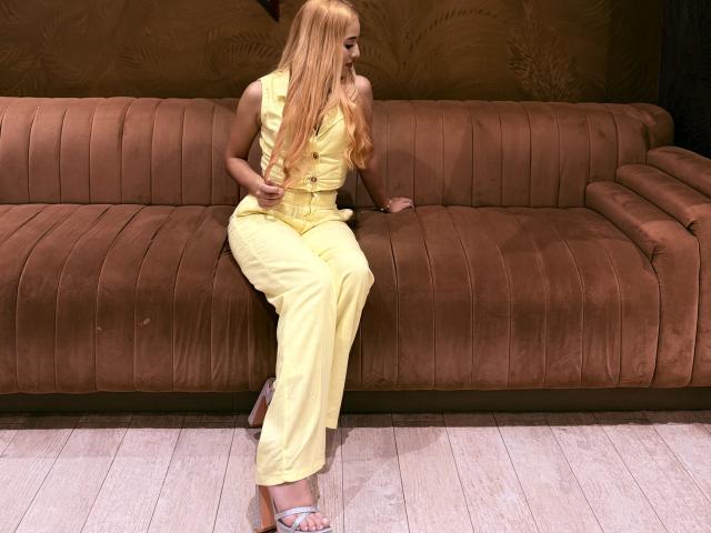EmmaWat with long blonde hair, yellow pantsuit, and white sandals, sitting on a brown suede couch, showing her ass, with a subtle smile, in a well-lit room.