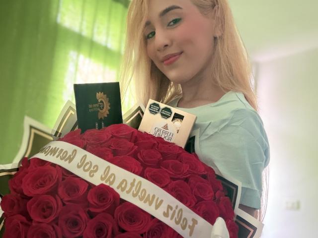 EmmaWat with blonde hair, blue shirt, holding red rose bouquet, smiling, green curtain background, small black and white cards in front, white ribbon on roses.