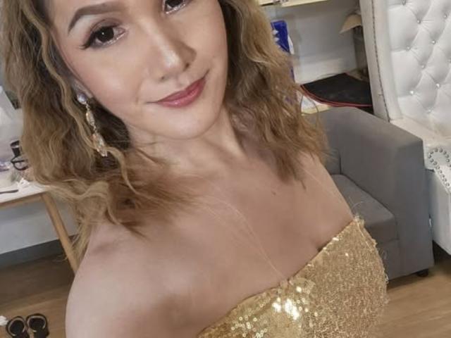 LovelyPinay69 with wavy blonde hair, wearing a sparkly gold strapless dress, showing her small tits, smiling at the camera, wearing gold earrings, in a cozy room.