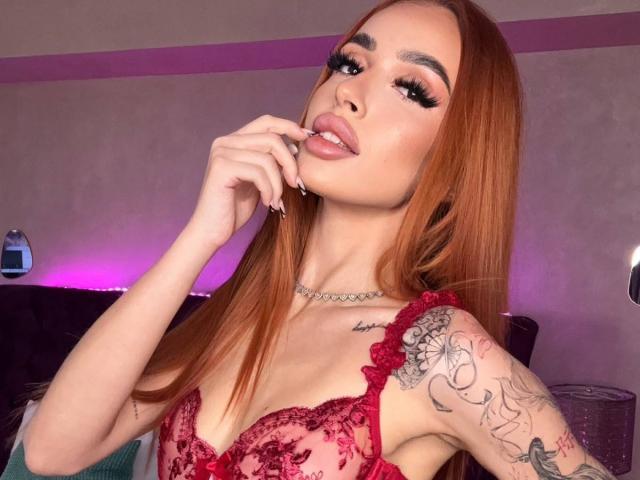 MarissaFex with red hair, wearing red lace lingerie, biting her finger, tattoos on her right arm, looking seductively at the camera. Her tits are visible through the lace.