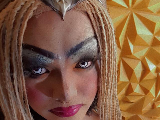 QueenOfPainSasha's face with blonde braids, dark gray eyeshadow, heavy eyebrows, pink lipstick, and intense blue eyes, wearing a metallic headpiece. Boobs are not visible.