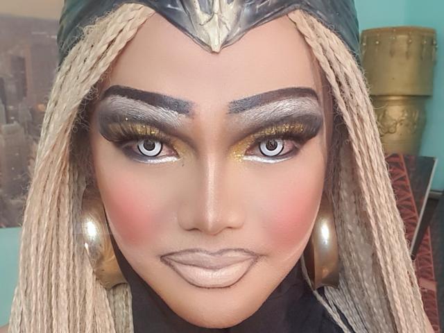 QueenOfPainSasha with tan skin, blonde braids, thick black eyebrows, gold eyeshadow, long lashes, shiny pink blush, metallic lips, gold headpiece, and large gold hoop earrings.