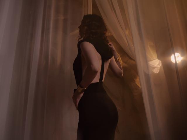 FabulousMagic with curly brown hair, wearing a black dress with suspenders, standing sideways, pulling up sheer curtains, showing a rounded ass, under warm, dim lighting.