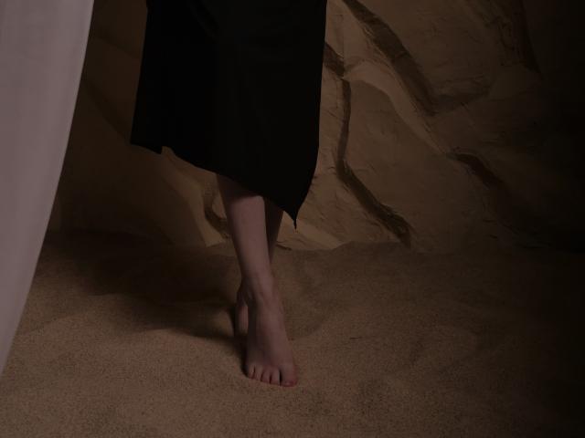 FabulousMagic's bare foot with pink toenails is in the sand, showing a slight toe curl, wearing a black skirt, in a dimly lit scene.