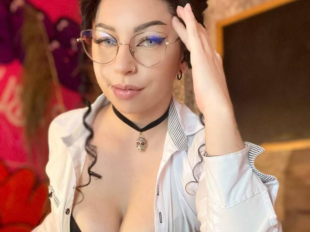 ScarlattaaDesire with glasses, black choker, and white shirt, touching her head, showing her tits, and a cute smile, has blue eyes and dark curly hair.