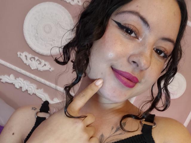 MiiaBennet has curly black hair, pink lipstick, and black eyeliner, with a finger pointing to her cheek. She has a shiny face and wears a black lace bra.