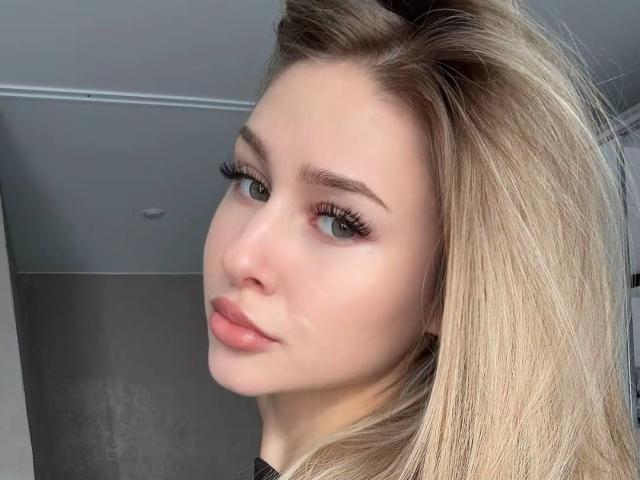 MilagrosEdmisten has blonde hair, green eyes, and full pink lips with long, thick eyelashes. Her fair skin is smooth, and she's looking to the side with a slight smile.