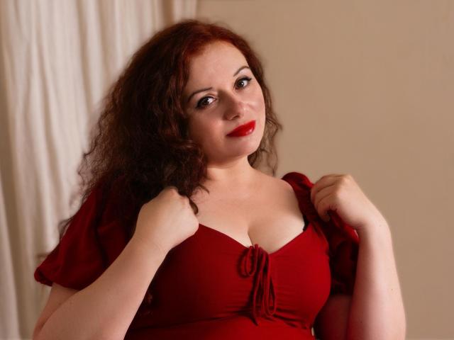 ElisabethLovee, com cabelo ruivo cacheado, batom vermelho e vestido vermelho, segurando as alças do vestido, mostrando os seios, sorrindo levemente, pele clara, corpo de porte médio, iluminação suave..