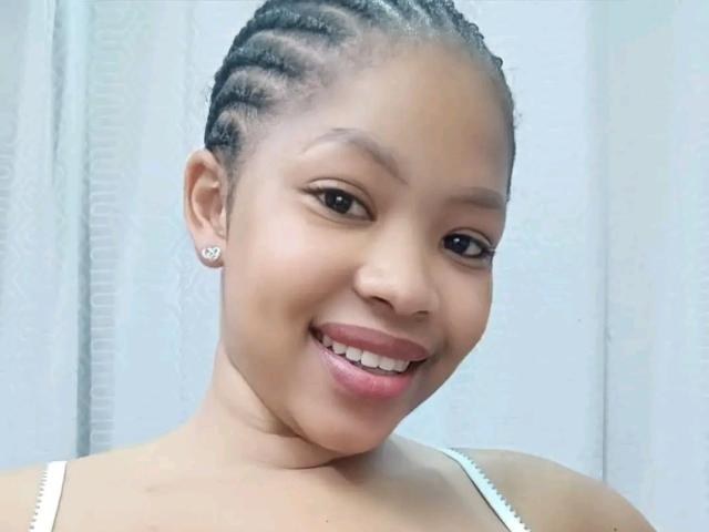 OnionBooty has medium brown skin, braided hair, small earrings, and is smiling with pink lips showing teeth. She's wearing a white strap top.