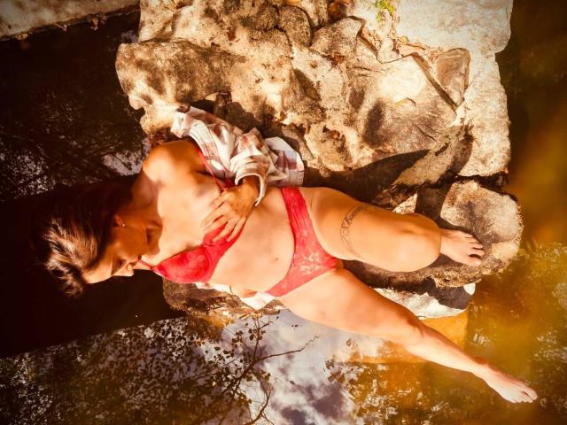 BKLove ligt op rotsen, gekleed in rode kant lingerie, bedekt haar borsten met één hand, haar billen zijn bloot, tatoeage op de rechter dij, in ondiep water, zonlicht werpt schaduwen..