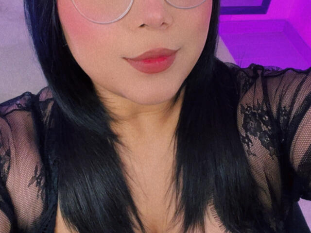 AlexaKiim with black lace top, showing her tits, pink blush, red lips, and black straight hair, wearing glasses, in a soft purple-lit room.