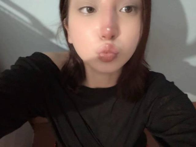 ElaneBoth puffs out her glossy, pink lips, making a kissing face with a slight upturn. She's wearing a black shirt, and her dark brown hair is shoulder-length.