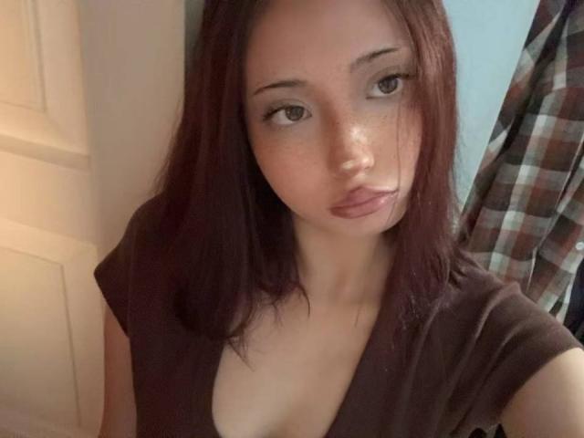 ElaneBoth has fair skin, dark red hair, and is wearing a brown top with a deep V-neck. She has freckles, full lips, and is looking slightly to the side.