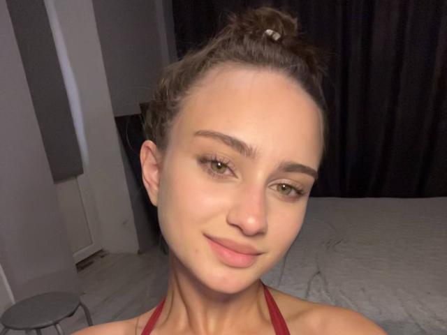 EleanorEvanchyk with brown hair in a bun, smiling, has a cute face, small nose, and is wearing a red halter top, showing her bare shoulders and a hint of her tits.