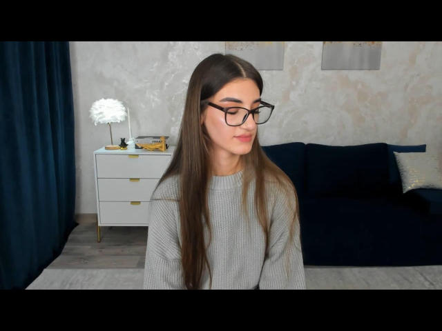 SabrinaWaynee with long brown hair, wearing black glasses and a gray sweater, sitting in a room with a dark blue couch and white dresser. She's looking down, smiling slightly.