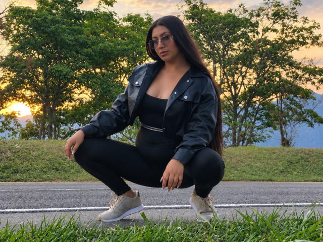 CatalinaRivero is squatting on a road in black clothes and a leather jacket, with long dark hair, glasses, and white sneakers, showing her ass and tits.