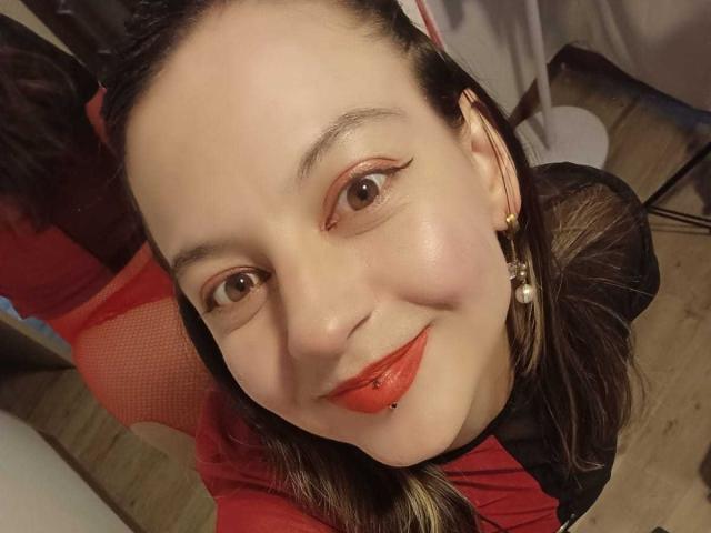 ArianaBlonde with fair skin, black hair, red lipstick, and a lip piercing, smiling at the camera, wearing a red shirt and pearl earrings.