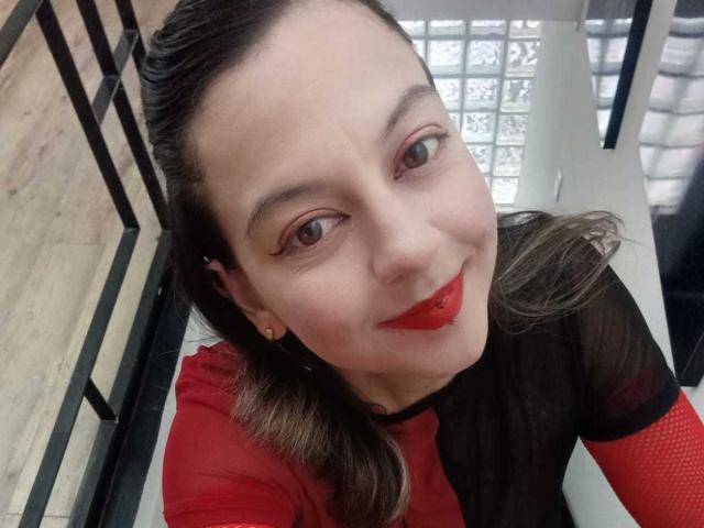 ArianaBlonde, with red lipstick, black mesh top, and red sleeves, smiling up at the camera. She has fair skin, brown eyes, and brown hair, wearing red lipstick.