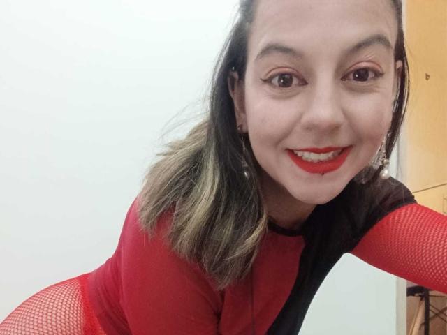 ArianaBlonde with red lipstick, smiling, wearing a red and black mesh bodysuit, leaning forward, showing her tits, brown eyes, and shoulder-length blonde hair.