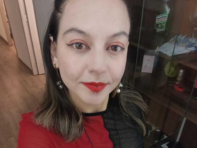 ArianaBlonde has red lipstick, pearl earrings, and black-red dress, with brown hair and subtle eyeliner, smiling at the camera. Her nose is pierced.