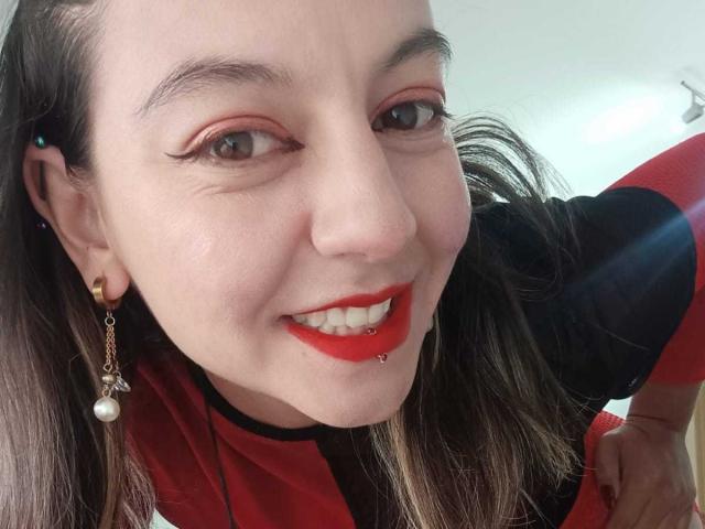 ArianaBlonde with red lipstick, pierced lower lip, and red eyeliner, smiling at the camera. She's wearing a black and red shirt, and has long brown hair.