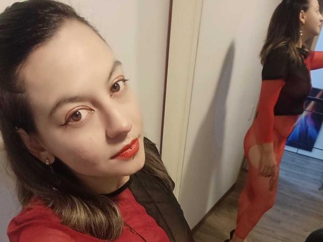 ArianaBlonde with red lipstick, wearing a red and black sheer top, showing her ass in a red mesh bodysuit, reflected in a mirror, standing in a wooden room.