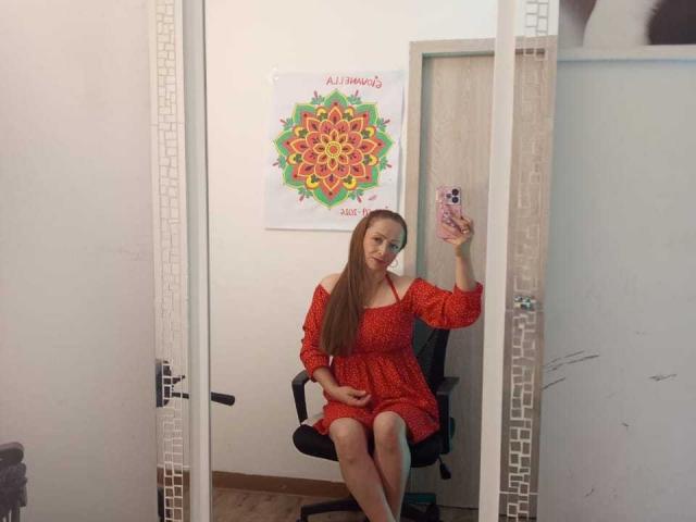 GianineLarose with long brown hair, wearing a red off-shoulder dress, sitting on a black chair, taking a mirror selfie, holding a pink phone, in a room with a mandala poster.