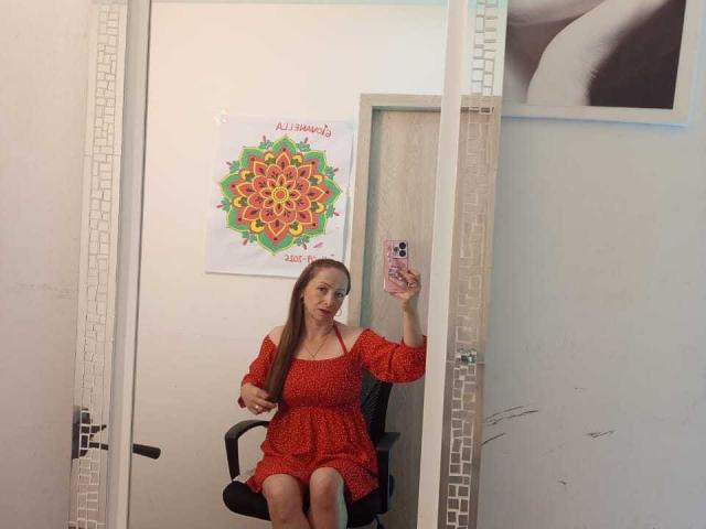 GianineLarose, con lunghi capelli castani, indossa un abito rosso con una spalla scoperta, è seduta su una sedia nera e sta scattando un selfie allo specchio con il suo telefono, mostrando i suoi piccoli seni..