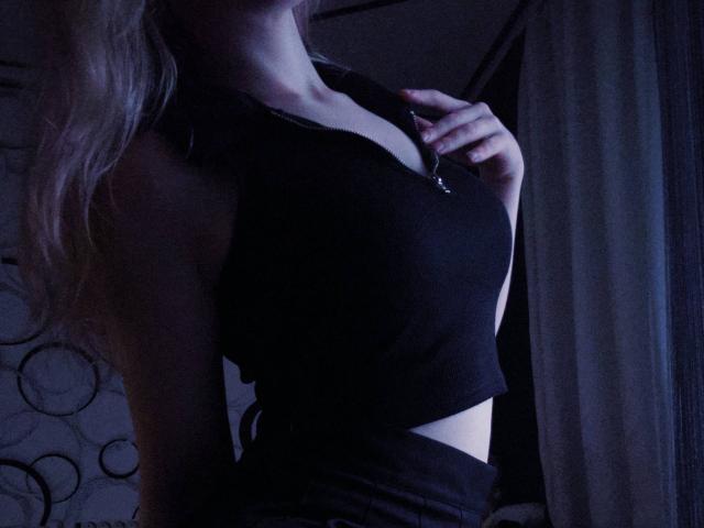 TiffaniMacandog's black crop top reveals her small tits, with her hand near her neck, in dim blue lighting. Her blonde hair is partially visible on the left.