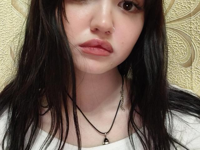 MistyEllstrom, con piel pálida, cabello negro y lacio, un piercing en la nariz y labios rosados, vestida con una camisa blanca y un collar negro, con una expresión seria. No se ven los senos..