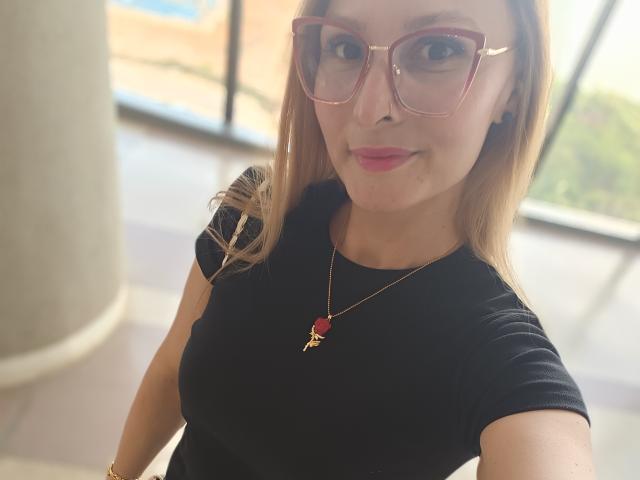 SachaClinford with blonde hair, pink glasses, black top, red pendant, small tits, smiling, pink lipstick, light skin, indoors, selfie, slight blur, casual, confident, cute.
