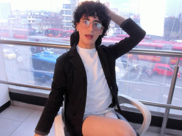 ElliotNoir, curly-haired with glasses, wearing a black jacket over a white tee and shorts, sitting with hand in curly hair, looking slightly open-mouthed, in a bright room.