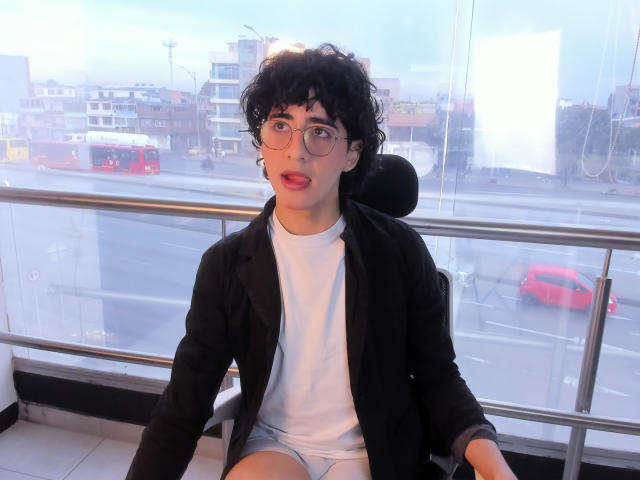 ElliotNoir with curly black hair, glasses, and a surprised expression, wearing a black jacket over a white t-shirt and blue shorts, sitting in a modern building.